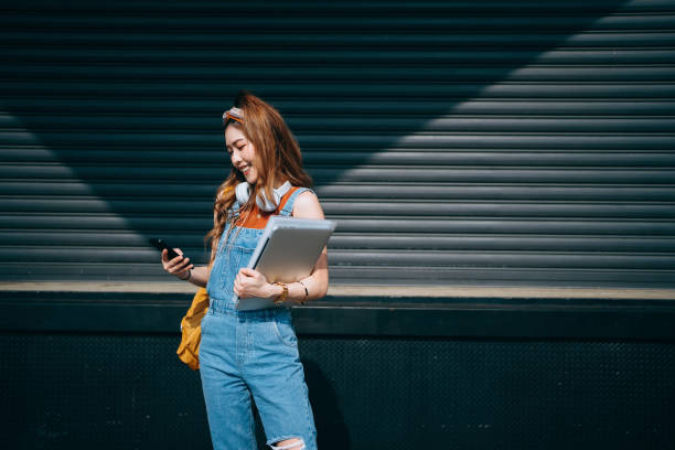 estudiante universitaria sonriente llevando mochila y usando auriculares alrededor de su cuello. ella está enviando mensajes de texto en el teléfono inteligente y sosteniendo la computadora portátil mientras camina en el campus de la escuela contra la l - back to school young women cheerful happiness fotografías e imágenes de stock