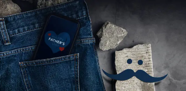 Fathers Day Concept. Text Message on Screen. Smartphone inside Jeans Pocket, surrounded by concrete pieces and Handmade Mustache