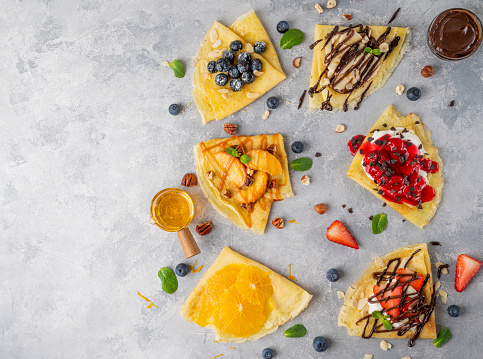 Variation of crepes or thin pancakes with fresh fruit, berries, cream cheese, honey, chocolate sauce on a gray concrete background. Top view, copy space.
