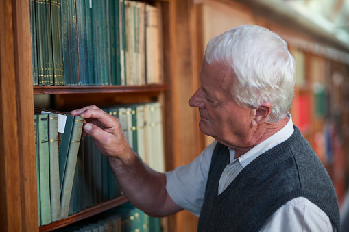 Portrait of mature man reading antique book in a big library. Active senior people. Smile on his face.