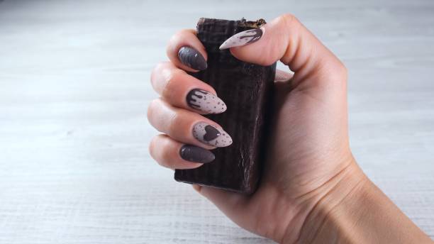 mains femelles avec de beaux ongles bruns de manucure sous forme de bonbons de chocolat, retenant une barre dépliée de chocolat noir, plan rapproché. profitez du dessert - brown chocolate candy bar close up photos et images de collection