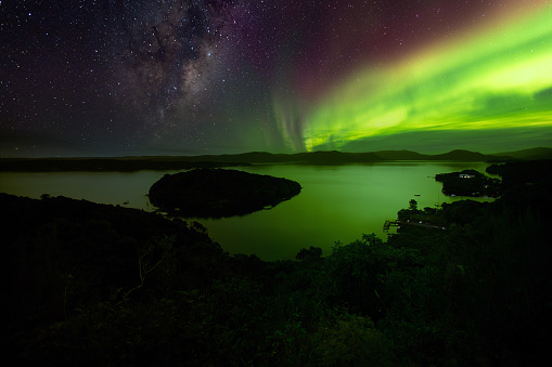 This composite image combines three images: A ground photo of Rakiura Stewart Island, a photo of the busy southern sky from Lake Tekapo, and an aurora photo from Norway. 

The ground image shows Paterson Inlet from the Observation Rock viewpoint in Oban. The northern lights (aurora borealis) were photographed from the village of Hamn. 

The southern lights (aurora australis) are sometimes visible from Rakiura Stewart Island. However, this photographer encountered overcast skies.