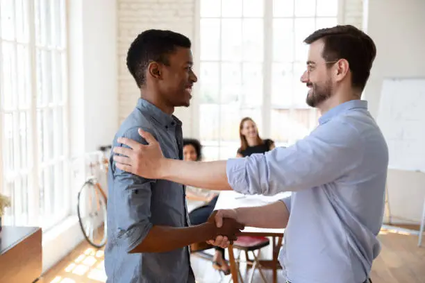 Photo of Smiling young male boss shaking hands with african employee.