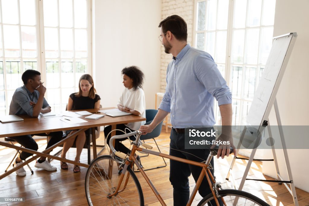 Creative diverse colleagues enjoying worktime in modern office. Young male manager came with bicycle, looking at mixed race diverse creative teammates discussing project ideas, solving problems sitting at table, eco-friendly healthy carbon free lifestyle concept. Corporate Business Stock Photo