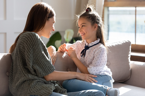 Happy young Caucasian mother and teen daughter sit relax on sofa talking sharing secrets. Smiling mom or nanny and teenage girl child rest on couch speak enjoy family weekend bonding at home.