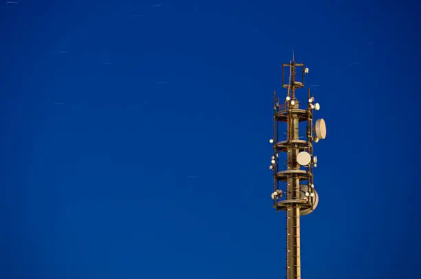 A telecommunications tower at night with blue sky and startrails.
