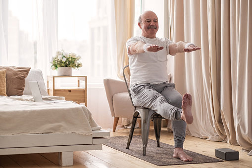 Elderly man practicing yoga asana or sport exercise for legs and hands using chair. Positive mood on sports activities.