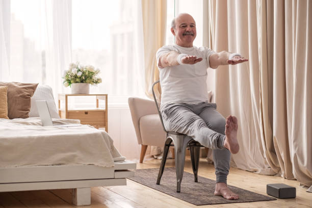 Elderly man practicing yoga asana or sport exercise for legs and hands on chair stock photo