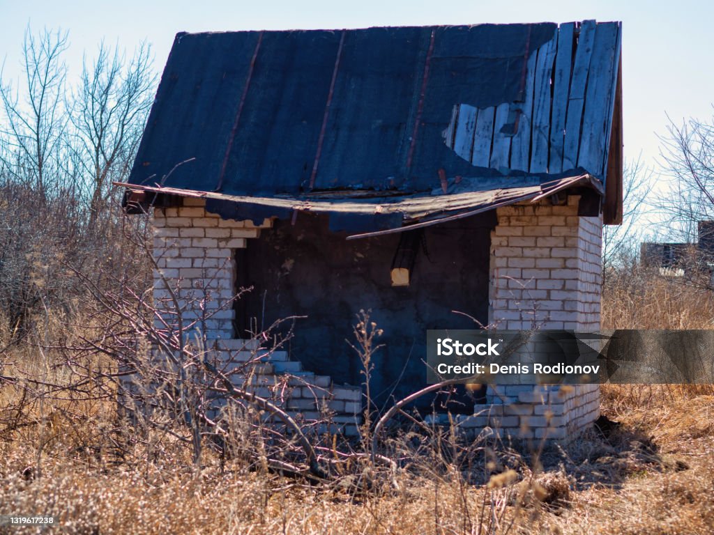 Broken brick house with a broken roof on a sunny spring day among the yellow thickets of grass. Broken brick house with a broken black roof on a sunny spring day among the yellow thickets of grass. House Stock Photo