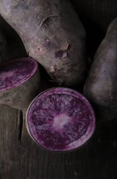 Close-up of sliced organic purple vitelotte truffle potatoes with skins and dirt on rustic wooden background. Also known as Truffe de Chine, Négresse, Vitelotte Noir or Black truffle potato.