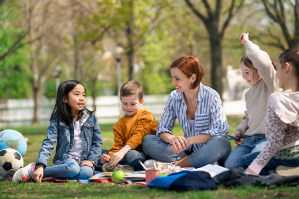 istock Teacher with small children sitting outdoors in city park, learning group education concept. 1319617766