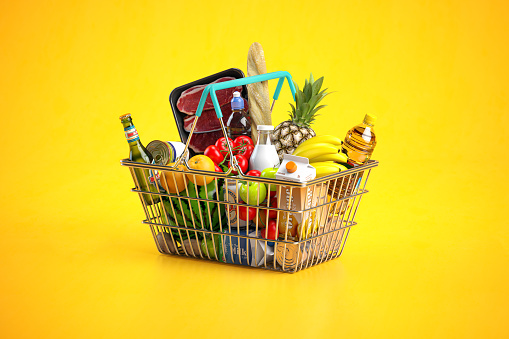 Shopping basket full of variety of grocery products, food and drink on yellow background.