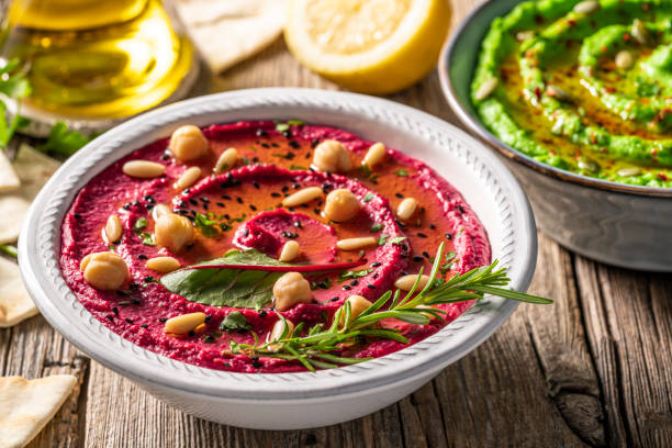 Beetroot pink hummus bowl closeup with pita flatbread dipping on rustic wood table garnished with black sesame and pine nuts