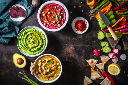 Hummus Three bowl of chickpeas, avocado and beetroot with cut vegetables for dip on dark wooden table background