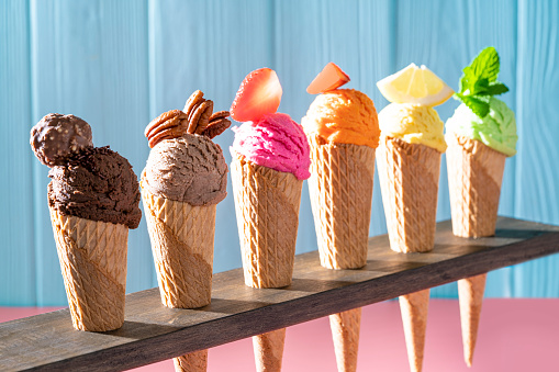 Assorted ice cream waffle cones in a row colorful different flavor as chocolate, mango, strawberry, mint, vanilla, lemon, coffee, nuts on pink and blue wooden wall
