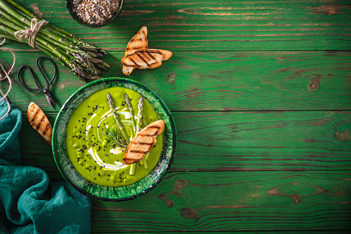 Asparagus soup plate with ingredients and bread toasts on green wooden table background