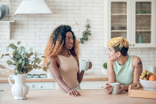 General interest. Two young adult concerned girlfriends in casual clothes communicating drinking coffee in kitchen at home