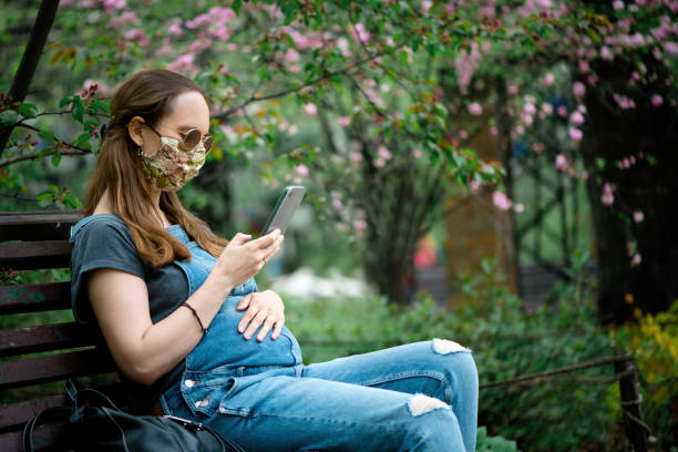 pregnant woman wearing protective face mask with smartphone in blooming park - using phone garden bench imagens e fotografias de stock