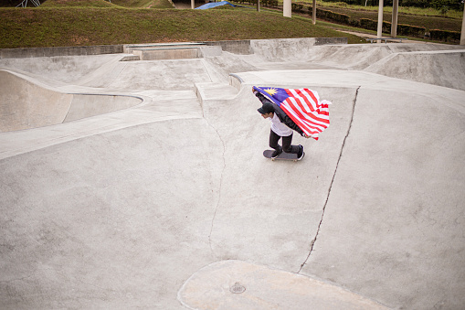 Asian skateboarder holding Malaysia flag riding skateboard in skatepark pool.