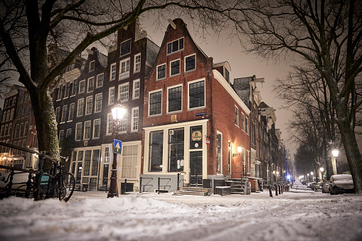 Street corner in the old part of Amsterdam during a night in winter. The streets are quite empty and covered in white from the snow. You can lightly see some snowfall. The street lightning is on just like the lights in the buildings. The sky is dark and trees stand tall next to the streets.