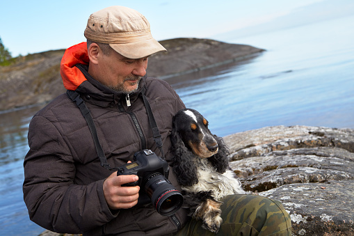Portrait of a middle-aged photographer and an English Cocker spaniel. A man sits on a stone ledge. He holds a camera in his right hand. A dog is sitting on the left. Lake Ladoga. Wildlife.