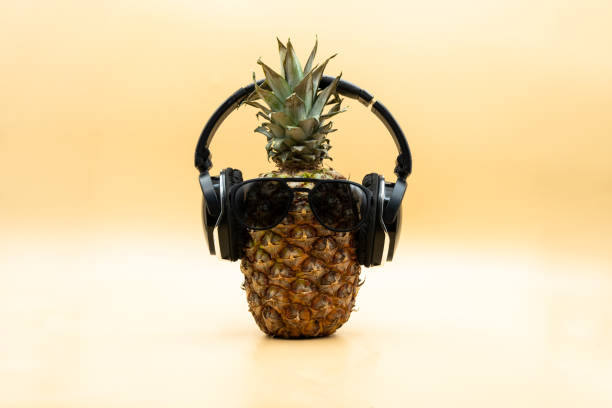 Funny pineapple in sunglasses wearing wireless headphones looking at camera over pastel yellow background stock photo