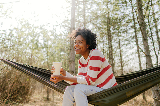 Photo of a young smiling woman relaxing in the hammock in the middle of the forest, sipping up a hot tea while enjoying in a beautiful autumn day she is spending outdoors.