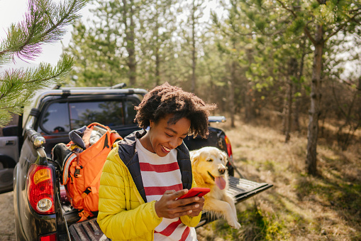 Photo of young smiling woman on a road trip with her best friend, her retriever dog, riding along on their pick up truck; using her mobile phone to check what she's missing on social medias