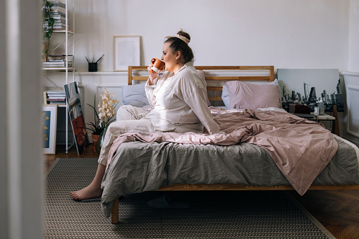 A young Caucasian plus size woman sitting on the bed in her pyjamas and having a cup of coffee in the morning