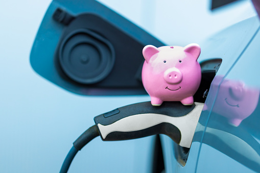 A pink piggy bank sits on the electric car's plug while it is charging. Concept of energy and electricity and saving money