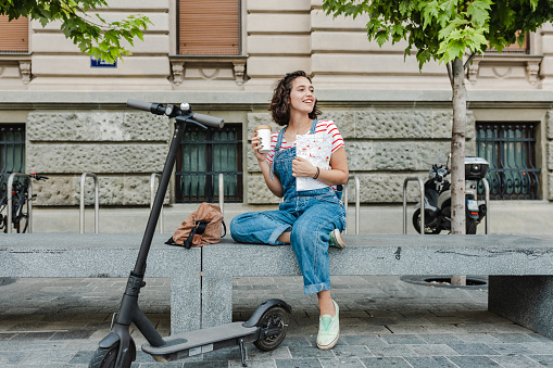 A woman with a backpack is relaxing and drinking coffee, she is leaning on an electric scooter
