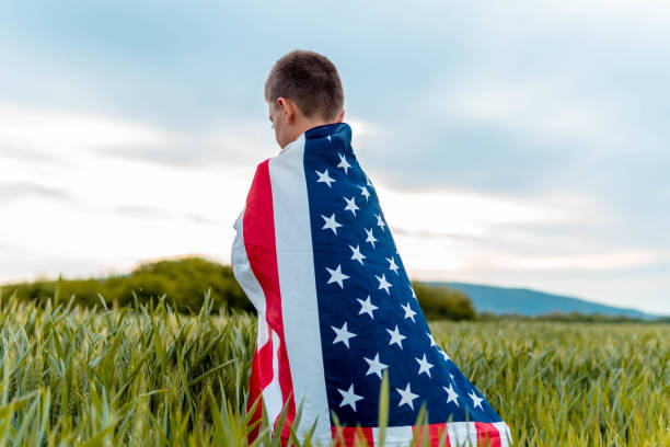 istock Sad boy wrapped with american flag. He is standing on a field looking sad and depressed 1319674219