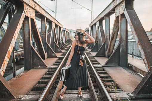 Young beautiful woman tourist in black dress on vacation wearing a hat standing on old rusty bridge