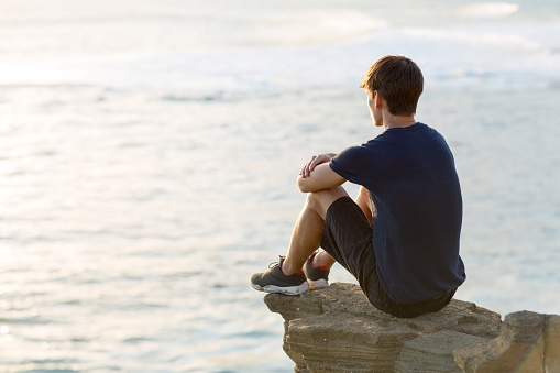 Thoughtful teen sitting alone on top of a cliff above the seaside view at dawn/sunset.