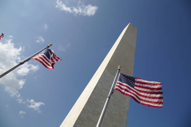 istock Two American flags in front of Washington Monument against blue sky with clouds. 1319685928