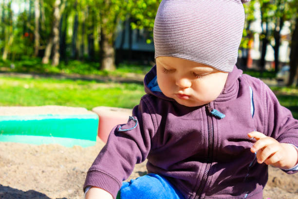 Cute little caucasian boy sitting in sandbox with toys on sunny summer day in park . Child looking at something in his hand, dressed in sweatshirt and striped hat.