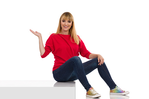 Casual young woman in red sweater is sitting on a step, looking at camera and presenting. Full length studio shot isolated on white.