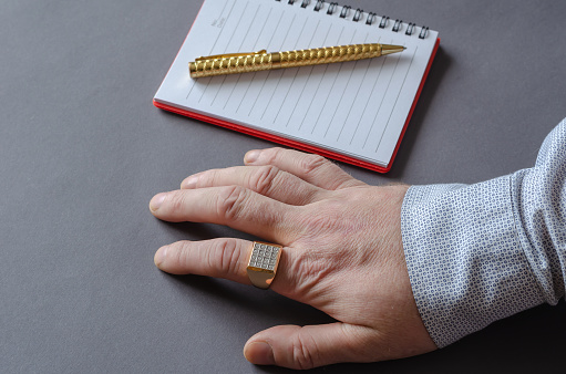 Hand, notepad, gold pen on gray background. Adult man with a seal on his index finger. Business concept.  View from above at an angle.