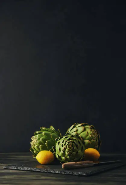 Still Life Background with Three Artichokes, Lemon and Kitchen Knife on a Slate Cutting Board