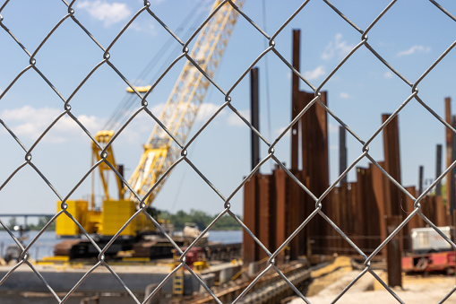 A chain link fence with a yellow construction crane and steel pilings at a waterfront site in a city during the day.