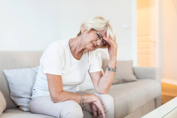 istock Mature woman sitting on a white sofa in a home touching her head with her hand while having a headache pain and feeling unwell 1319719991