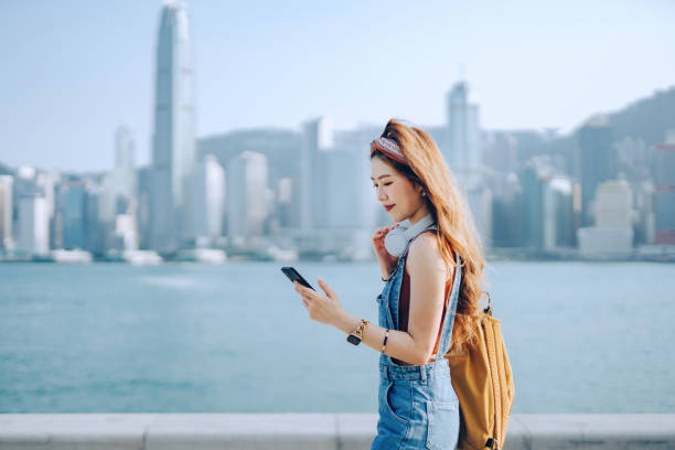 hermosa joven asiática llevando mochila y usando auriculares alrededor de su cuello. ella está enviando mensajes de texto en el teléfono inteligente mientras camina a lo largo del paseo marítimo contra el espectacular horizonte urbano de la ciudad en u - nativo digital fotografías e imágenes de stock
