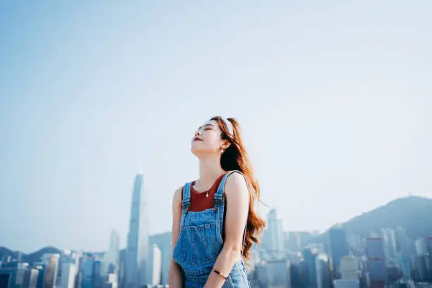 Photo of Beautiful smiling young Asian woman closed her eyes while taking a deep breath, enjoying the fresh air and the gentle breeze outdoors, against spectacular urban city skyline by the promenade of Victoria harbour