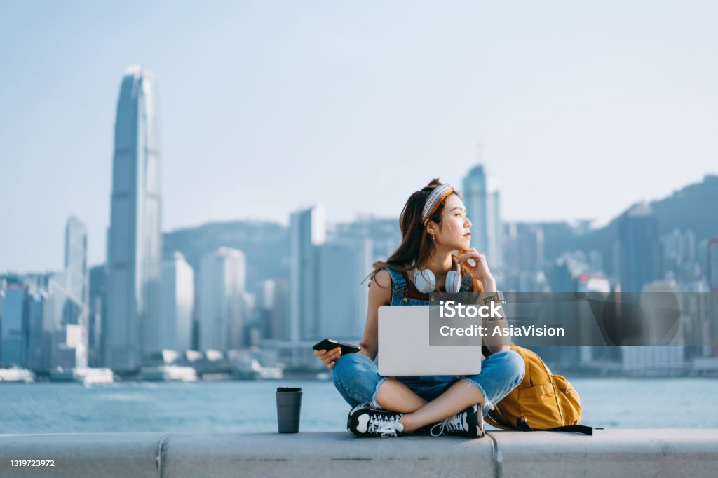 Beautiful young Asian woman sitting cross-legged by the promenade, against urban city skyline. She is wearing headphones around neck, using smartphone and working on laptop, with a coffee cup by her side. Looking away in thought. Lifestyle and technology Asian and Indian Ethnicities Stock Photo