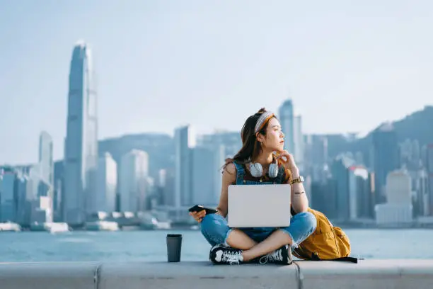 Photo of Beautiful young Asian woman sitting cross-legged by the promenade, against urban city skyline. She is wearing headphones around neck, using smartphone and working on laptop, with a coffee cup by her side. Looking away in thought. Lifestyle and technology