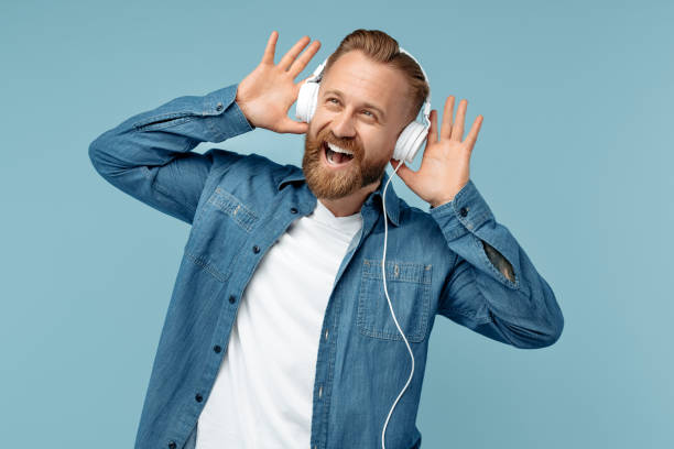 青い背景の上に白いスタイリッシュなヘッドフォンで音楽を聴くジーンズシャツで幸せなひげを生やしたブロンドのヒップスターの男。 - free standing audio ストックフォトと画像