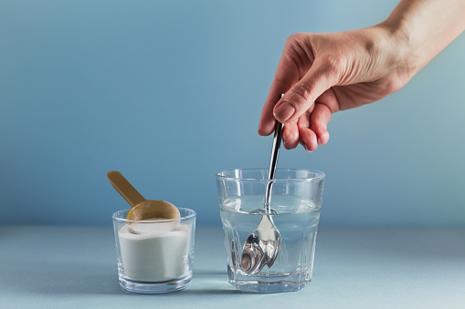 Glass with collagen dissolved in water and collagen protein powder on light blue background. Woman's hand holds a spoon. Healthy lifestyle concept.