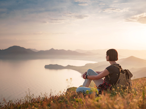 A young Asian woman with a backpack Sitting on a hill slope and enjoying the view of the sunset sea and mountains . Mountain and coastal travel, freedom and an active lifestyle