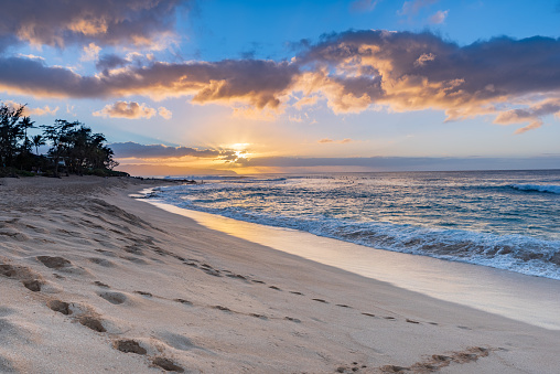 View of Sunset Beach on the North Shore of Oahu, Hawaii at sunset with palm trees, beach, and waves rolling in over rocks in the foreground