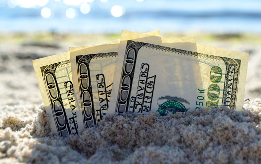 Three dollar bills are buried in sand on sandy beach near sea on sunny bright summer day close-up. Dollar bills partially buried in sand